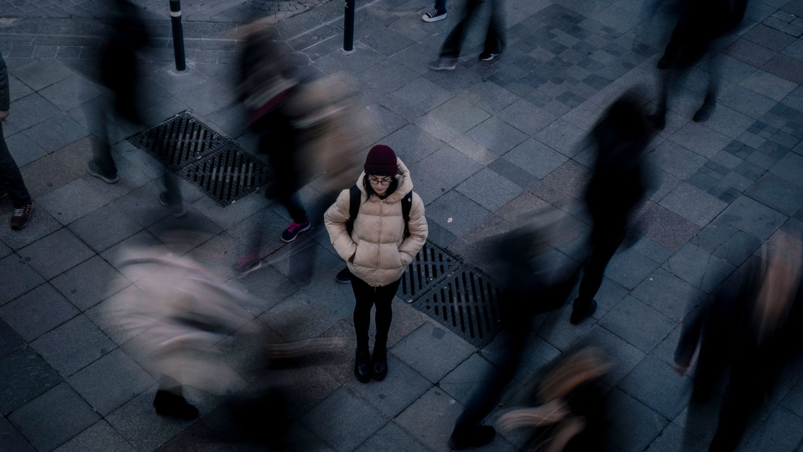 A still woman standing as a blurred crowd of people streams past her on a city street.