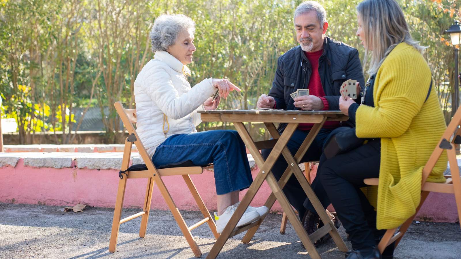 Three older friends playing cards together at a table outdoors in a park.