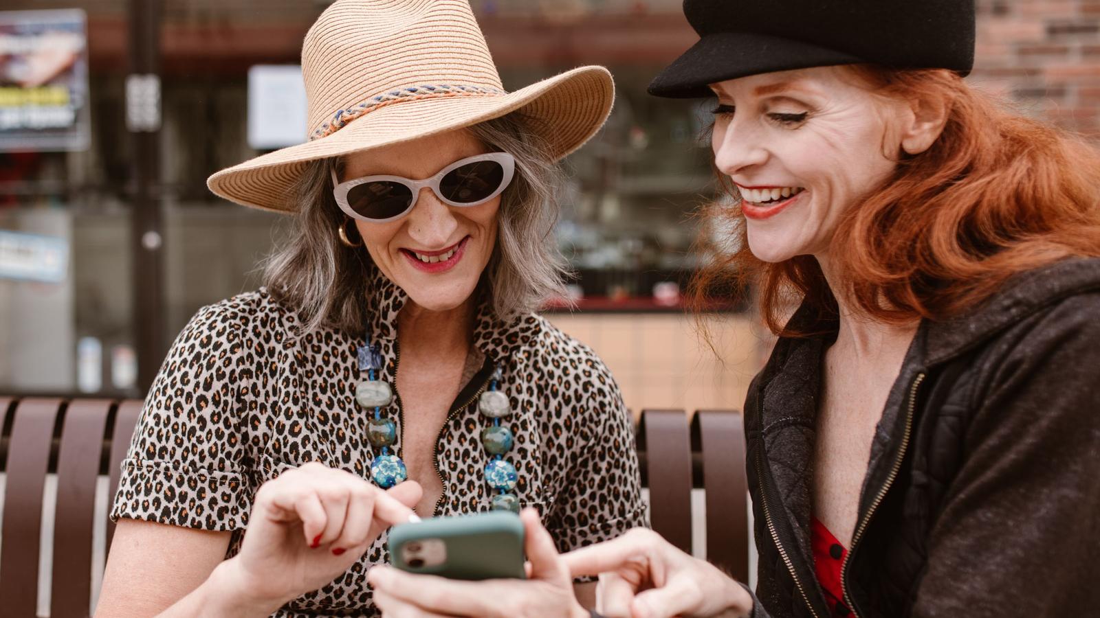 Two women in their fifties laughing together over a phone outdoors, an easy moment of friendship.