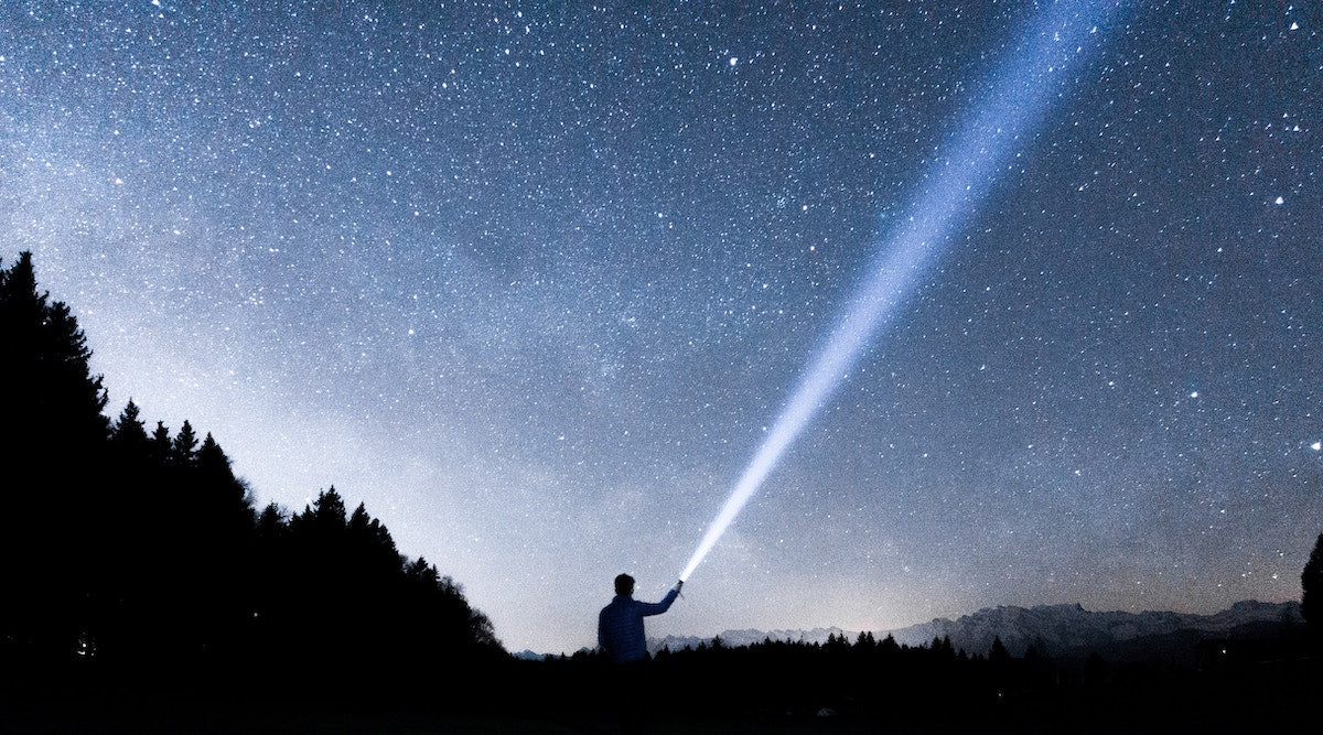 Silhouette of a person shining a flashlight toward a starry night sky — per aspera ad astra, through hardships to the stars